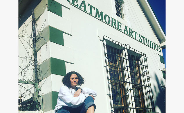 Woman with brown curly hair, wearing a white jumper smiles and sits below a white and green building that reads Greatmore Art Studios.