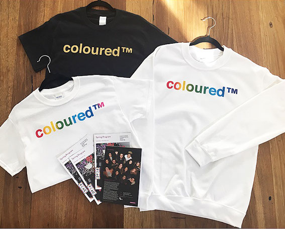 A collection of tshirts and sweater, white with rainbow text, black tee with gold text that all have the words printed colouredtm upon them.