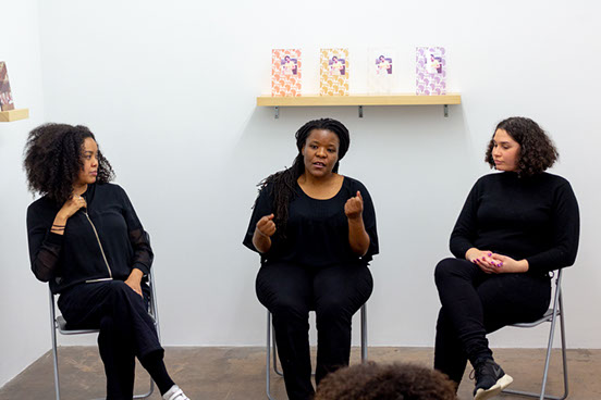 Three African diaspora women sitting on chairs, wearing all black clothes in a white gallery space with artworks behind them.