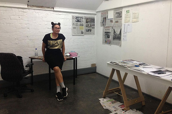 Woman wearing black and gold top and black shirt leaning on a desk in an artist studio with newspaper clippings on wall, trestle table and floor